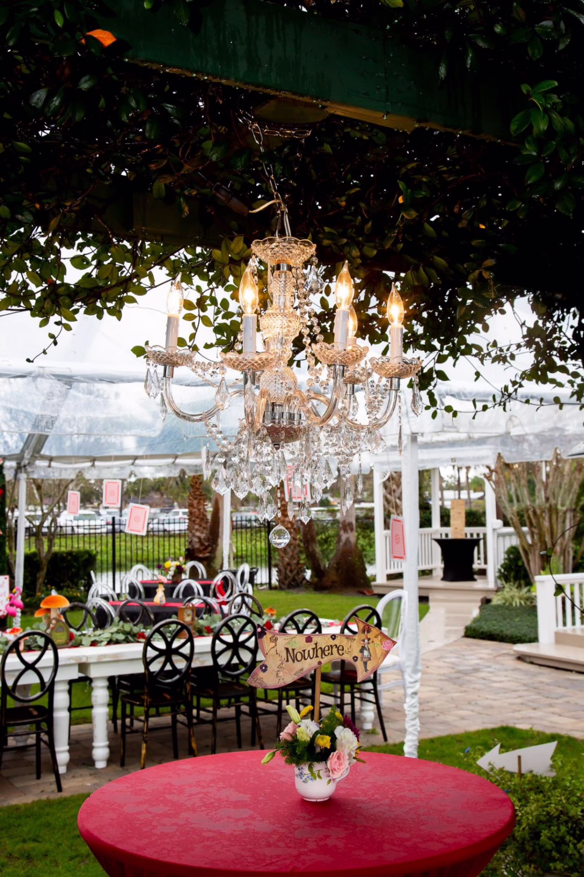 alice in wonderland themed party setup orlando celebration gardens winter park - Pricing Alice in Wonderland themed party setup with crystal chandelier hanging from pergola, red round table with floral centerpiece and Nowhere directional sign at Orlando Celebration Gardens wedding venue in Winter Park Florida