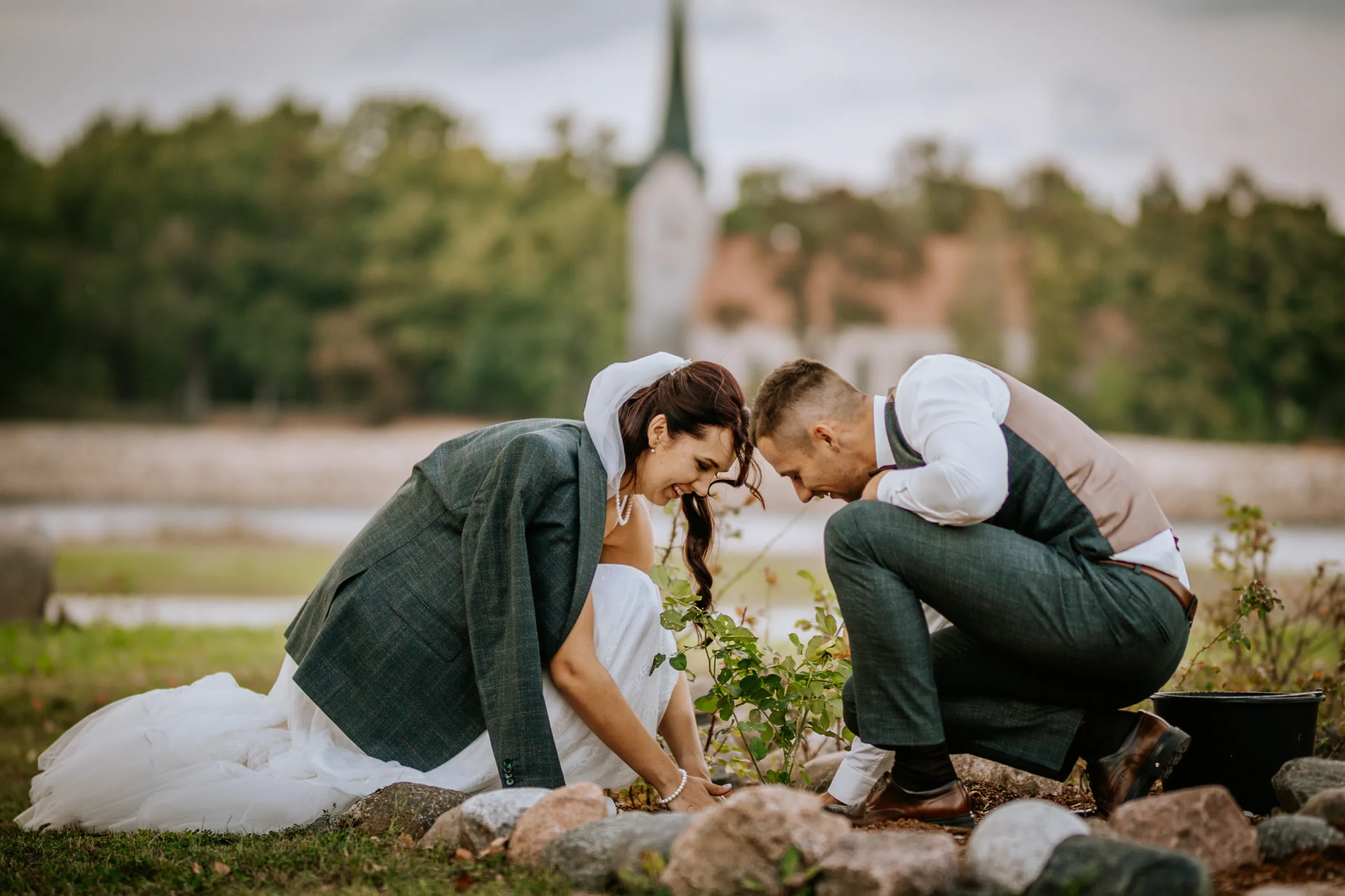 Smiling bride and groom in wedding attire planting a tree together outdoors near a church steeple, a meaningful wedding day unity tradition