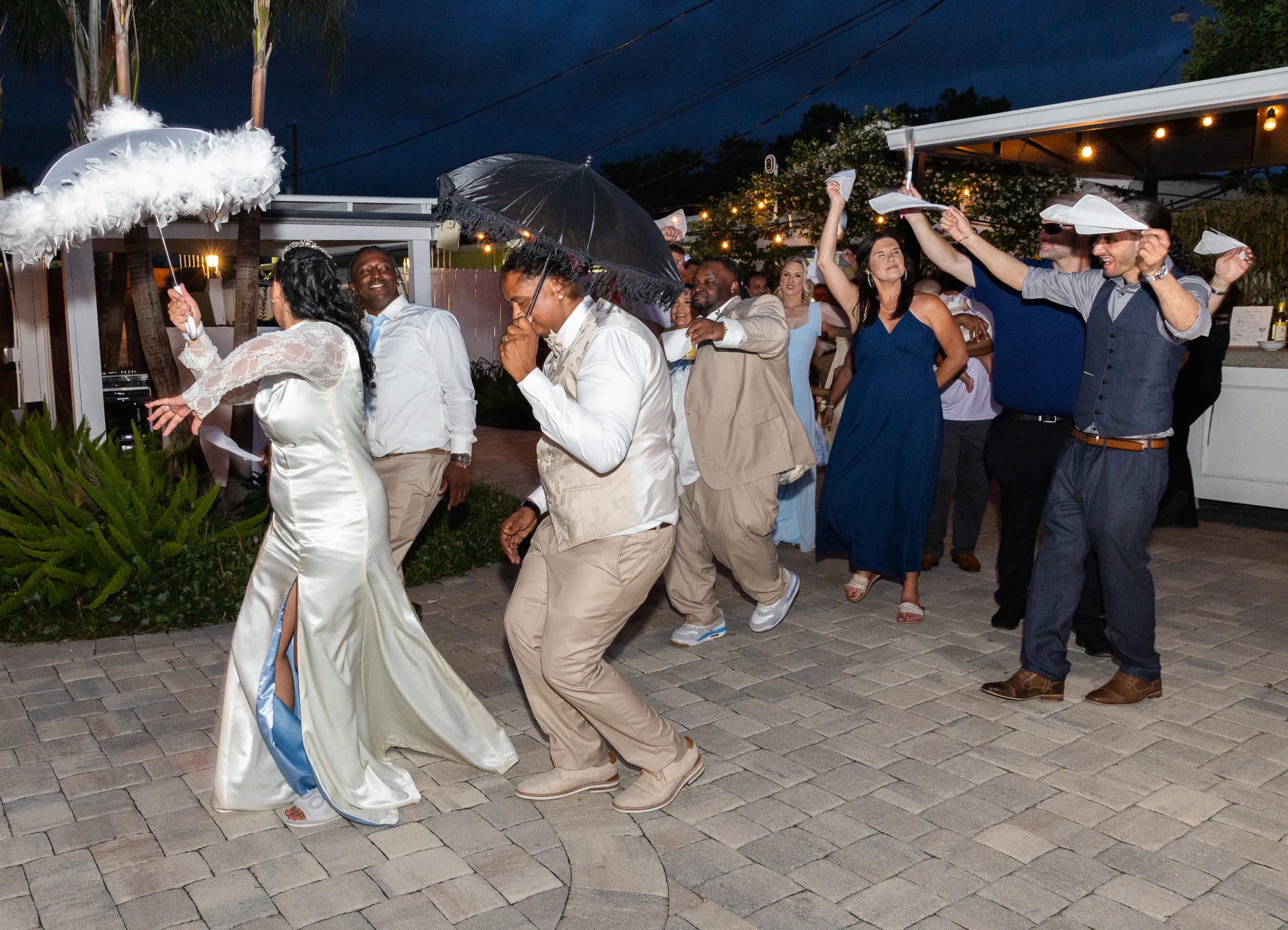Bride and wedding party perform a traditional second line parade dance with feathered parasols and umbrellas during outdoor evening wedding reception in Orlando FL