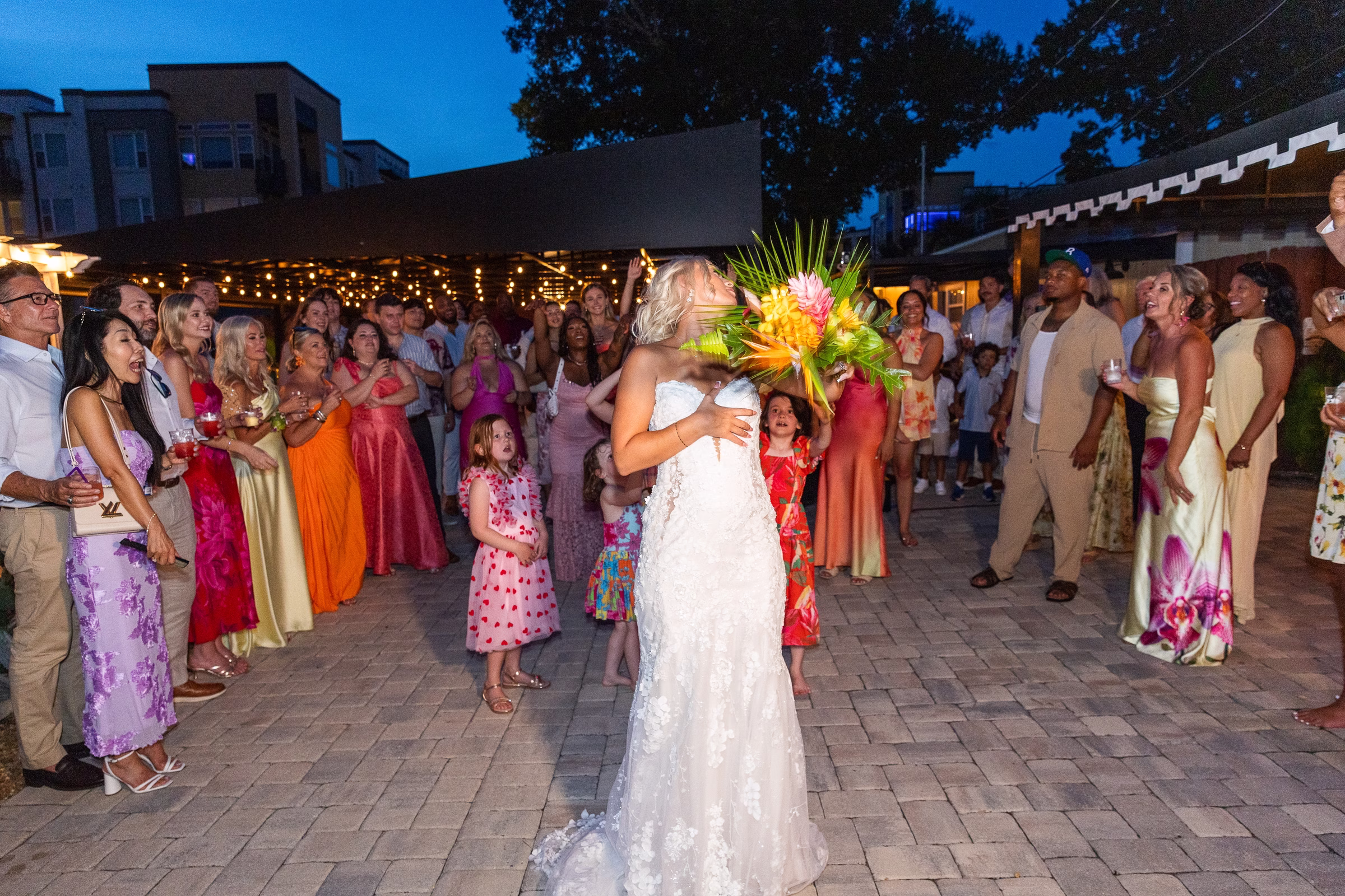 Bride in a lace wedding gown tosses a vibrant tropical bouquet to excited guests during an outdoor evening reception at Celebration Gardens wedding venue in Orlando Florida