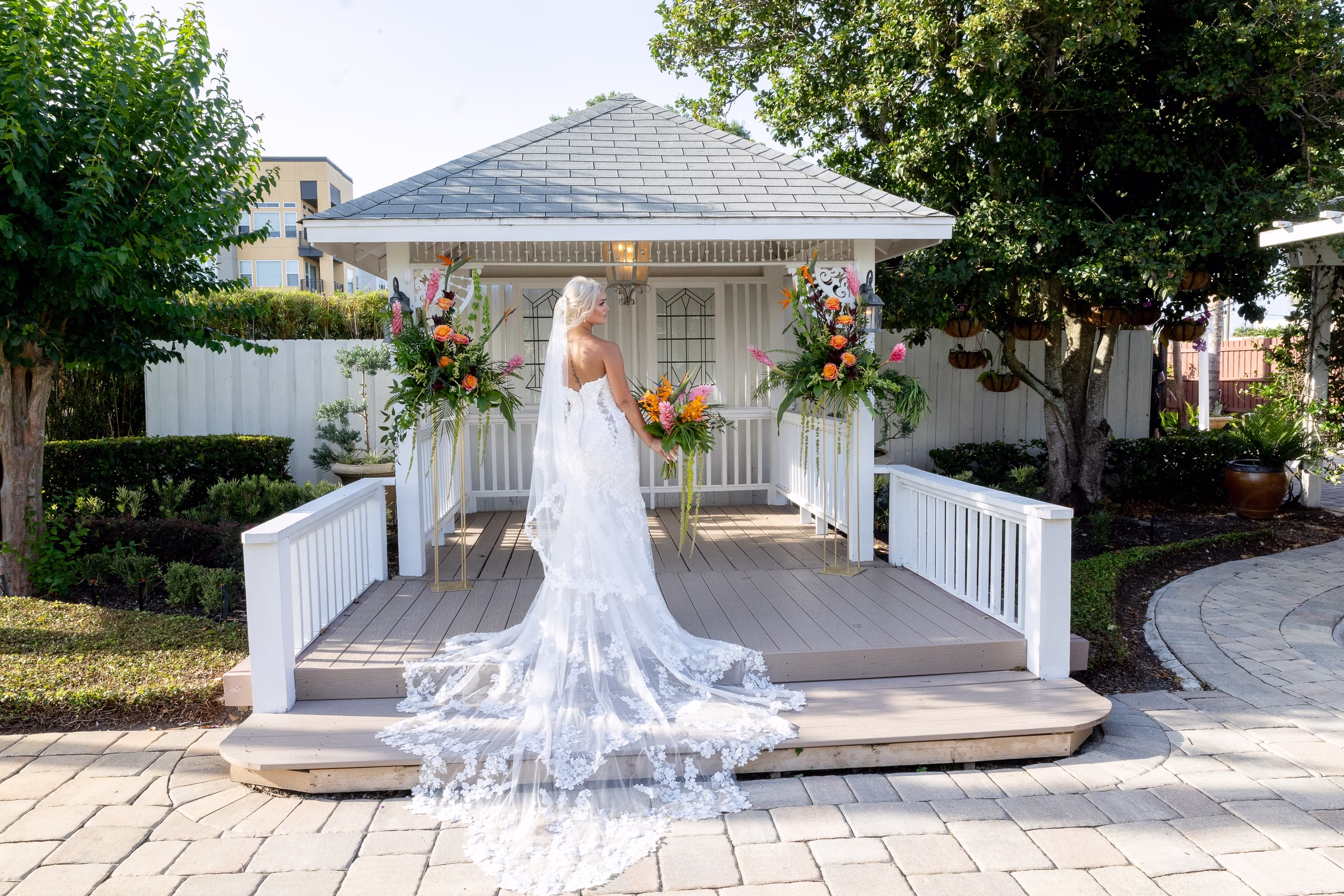 Bride in a lace gown with cathedral veil and train poses on the gazebo deck at Orlando Celebration Gardens wedding venue in Orlando Florida surrounded by vibrant tropical floral arrangements
