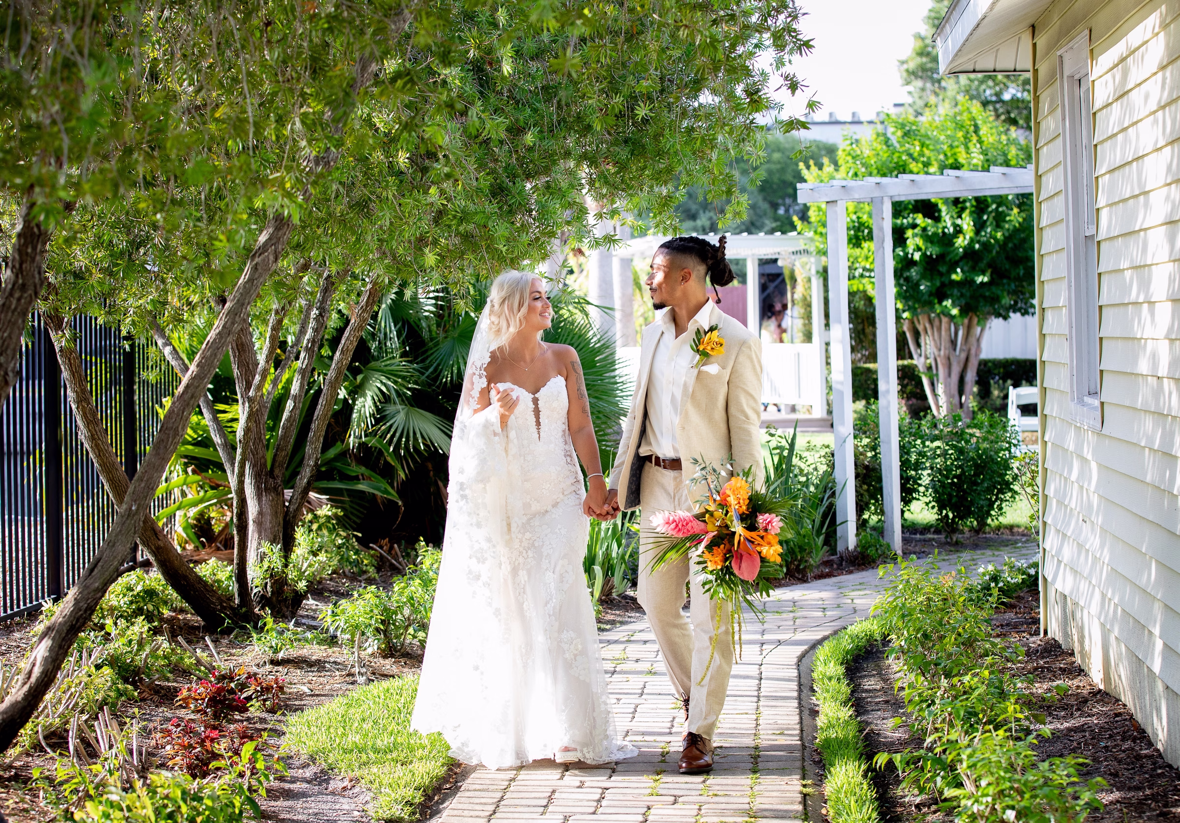 Bride in lace gown and groom in tan suit holding hands and walking along a brick garden path at Orlando Celebration Gardens wedding venue in Orlando Florida