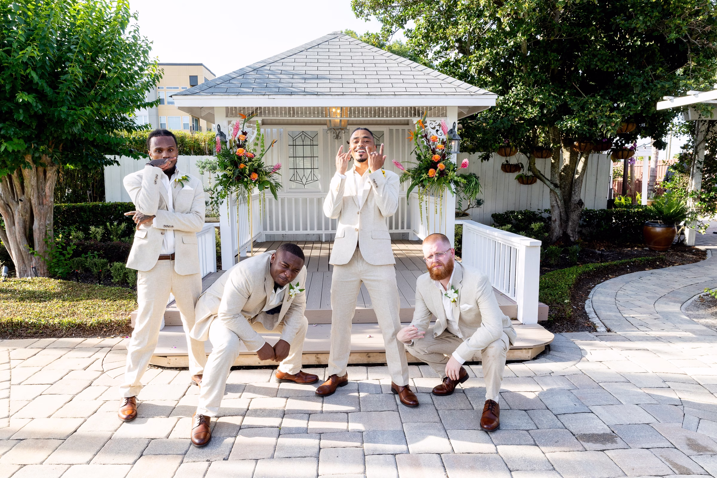 Groom and groomsmen in cream suits striking fun poses in front of the floral-decorated gazebo at Orlando Celebration Gardens wedding venue in Orlando Florida