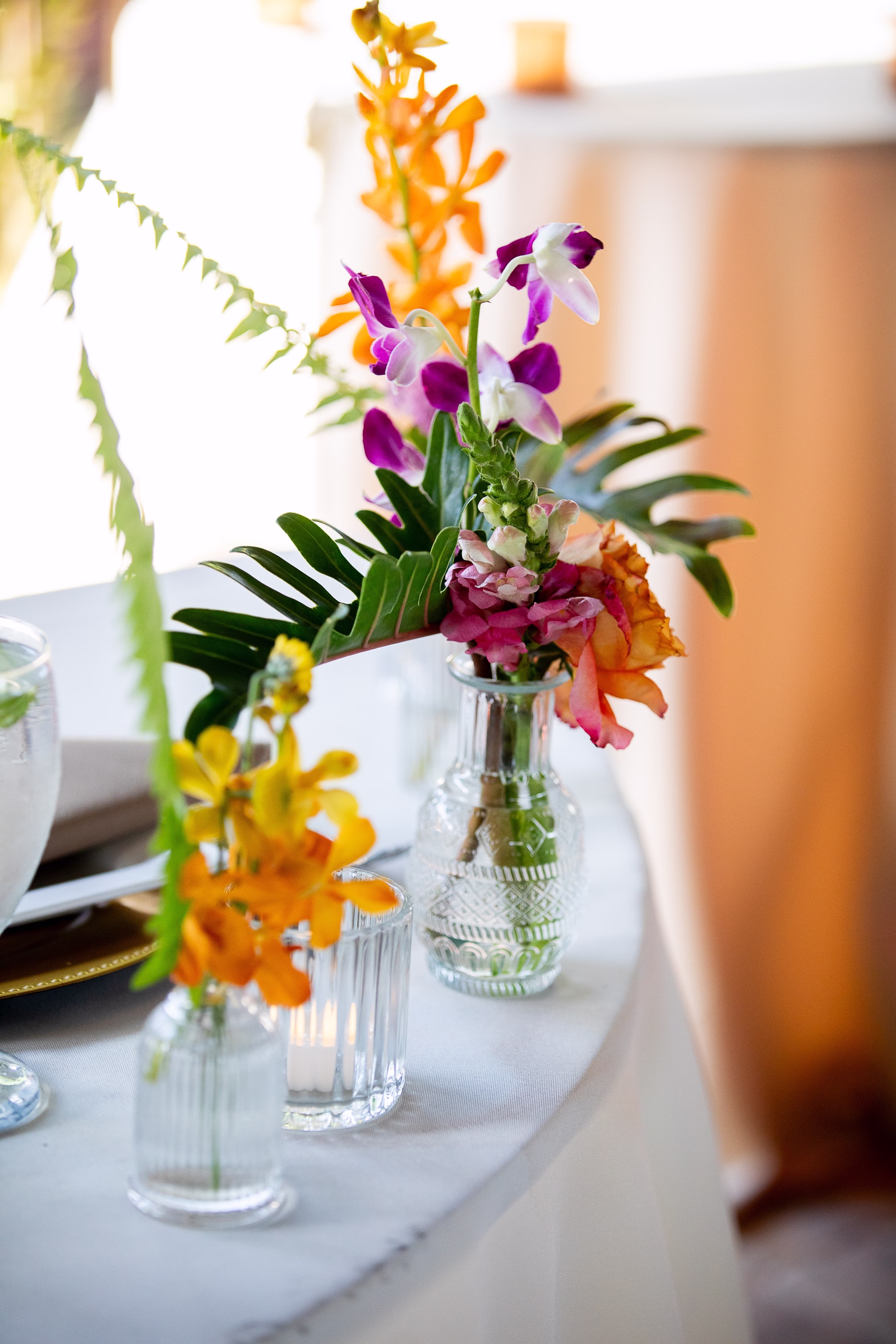 Colorful tropical floral centerpiece with purple orchids, orange blooms, and monstera leaves in glass bud vases on a white linen table at Orlando Celebration Gardens wedding venue in Orlando Florida