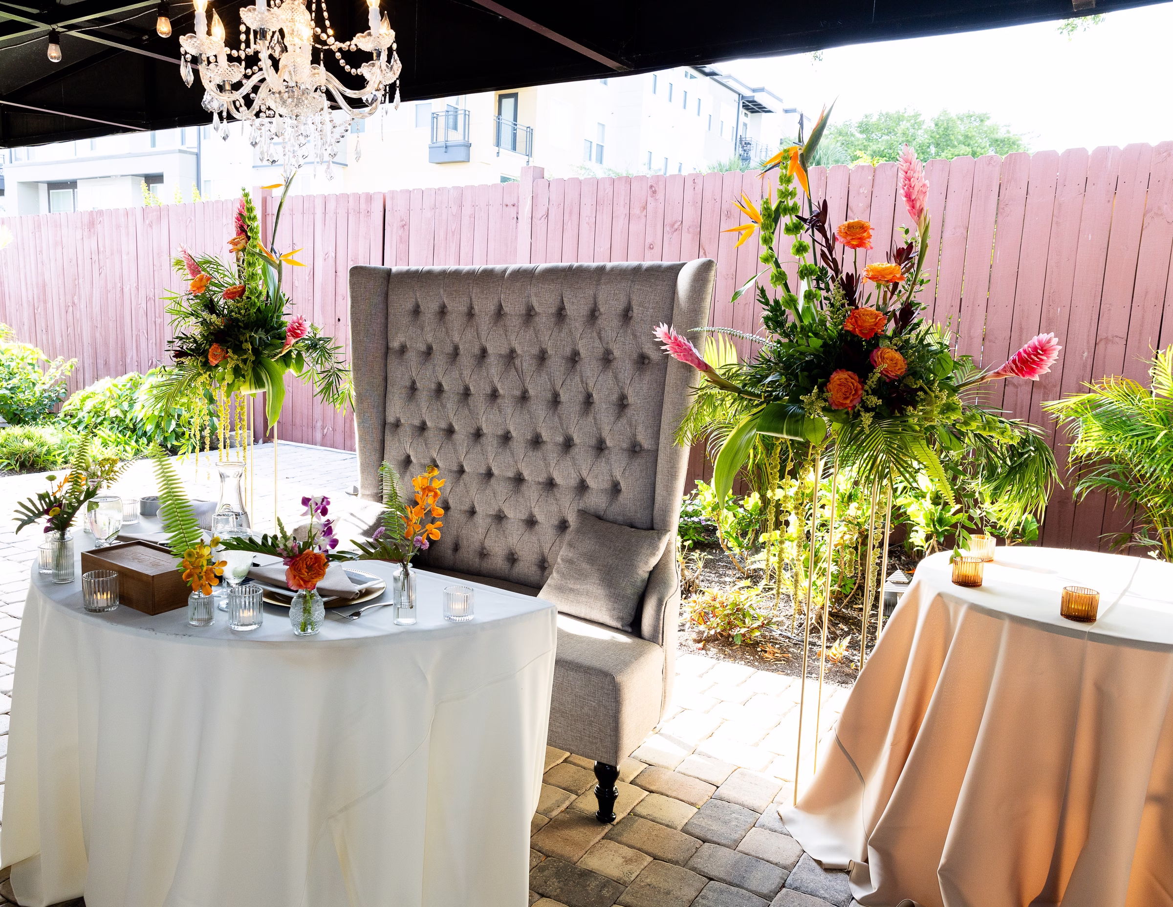 Tropical floral arrangements with bird of paradise and orange roses flanking a tufted wingback sweetheart chair at Orlando Celebration Gardens outdoor patio wedding venue in Orlando Florida