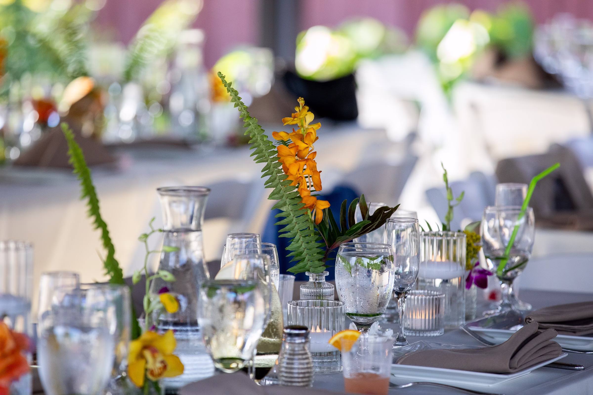 Elegant tropical wedding reception tablescape featuring orange mokara orchids, fern fronds, crystal glassware, and candlelight at Orlando Celebration Gardens in Orlando Florida