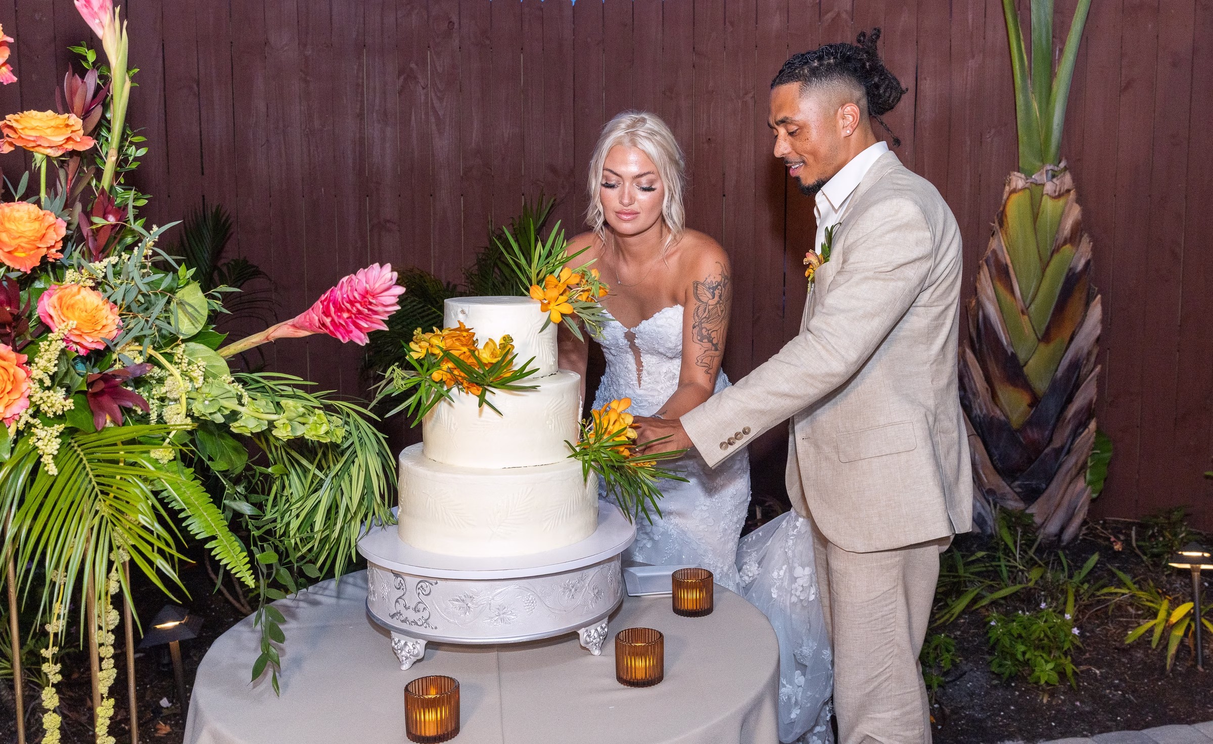 Bride and groom cutting a three-tier white wedding cake adorned with tropical orange and yellow flowers at Orlando Celebration Gardens in Orlando Florida
